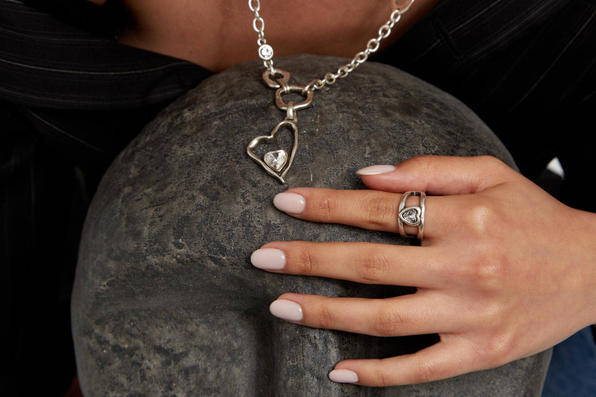 A silver heart-shaped pendant necklace with a clear heart gemstone, shown on a dark gray mannequin head with a person's hand resting on it. The person is wearing a silver ring with a matching heart gemstone.