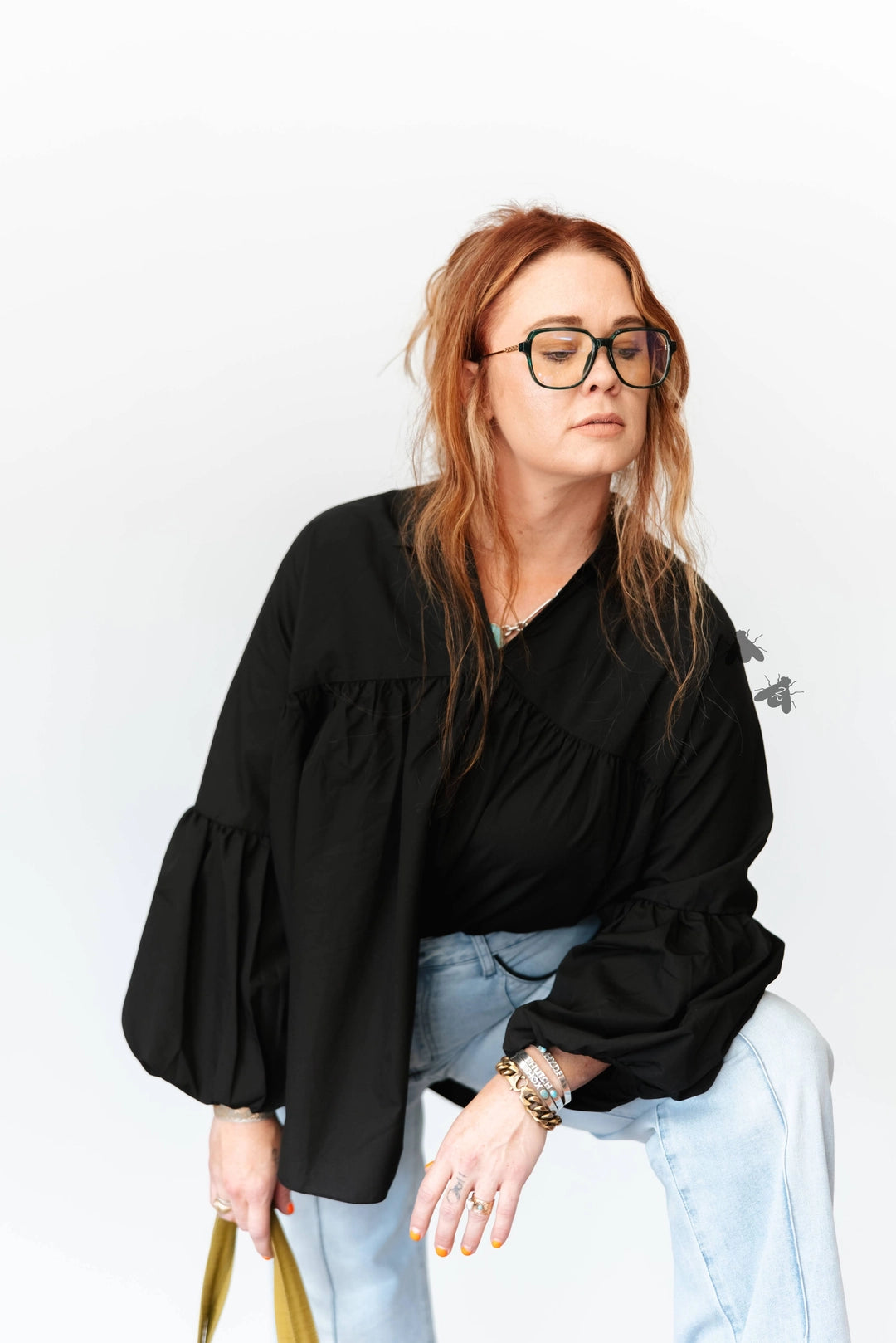 A woman with red hair and glasses wearing a black long-sleeve blouse with dramatic ruffled sleeves, paired with light blue jeans, posing against a white studio background.