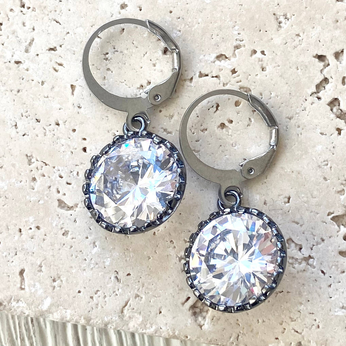 A pair of silver-toned leverback hoop earrings with large, round, clear faceted gemstones in a beaded silver border, displayed on a light beige textured stone surface.