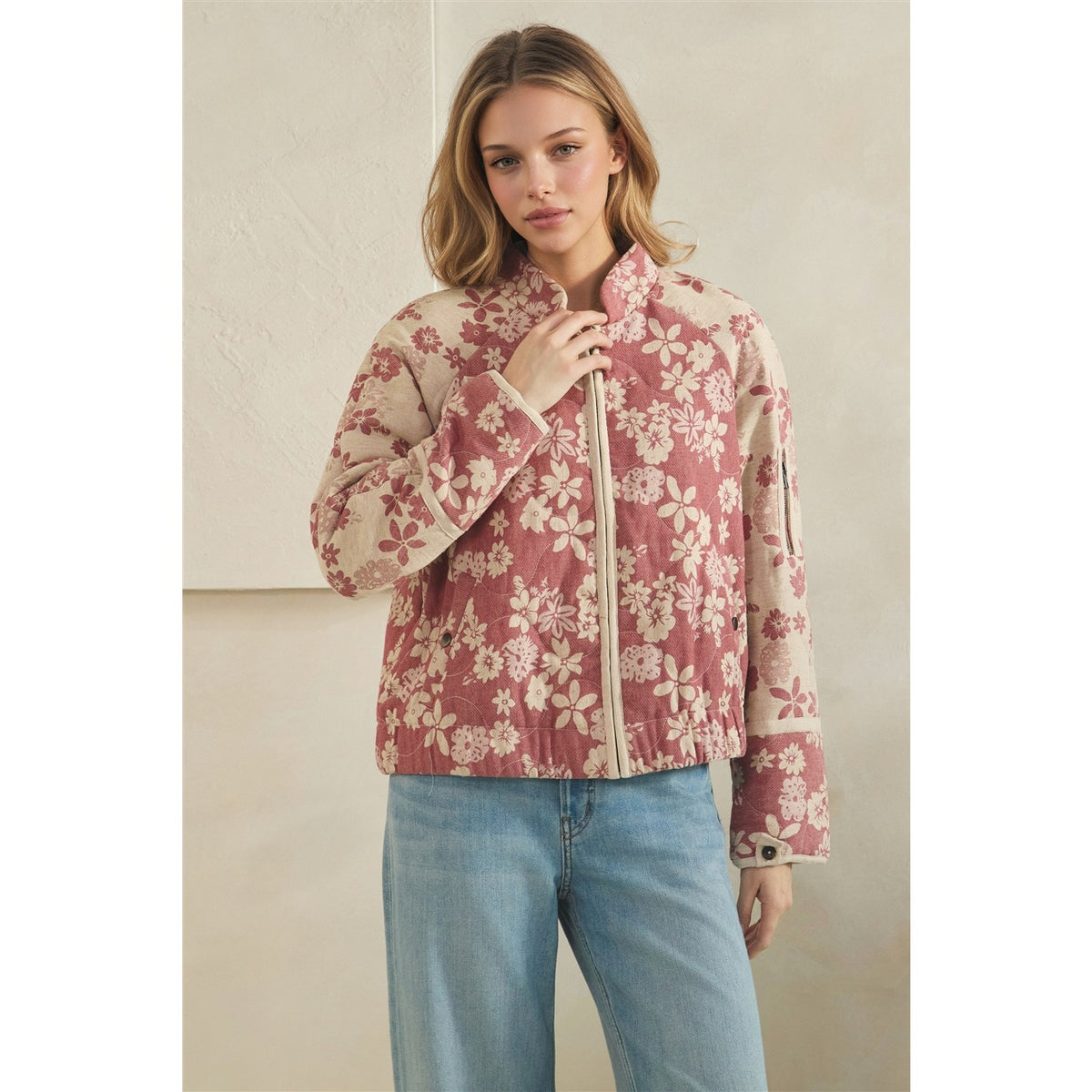 A woman wearing a cropped quilted jacket with a dusty rose and cream floral pattern, paired with light blue jeans, standing against a neutral beige wall.