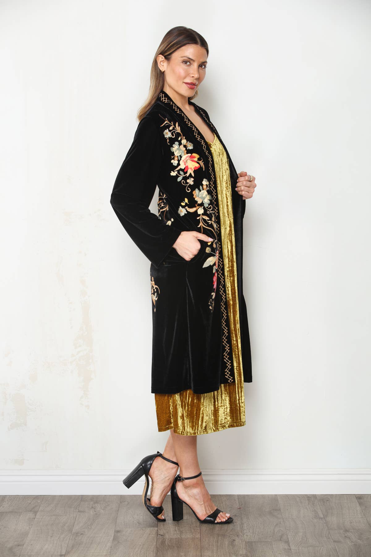 Angled front view of black floral embroidered velvet duster with model’s hand in side pocket over a gold dress.