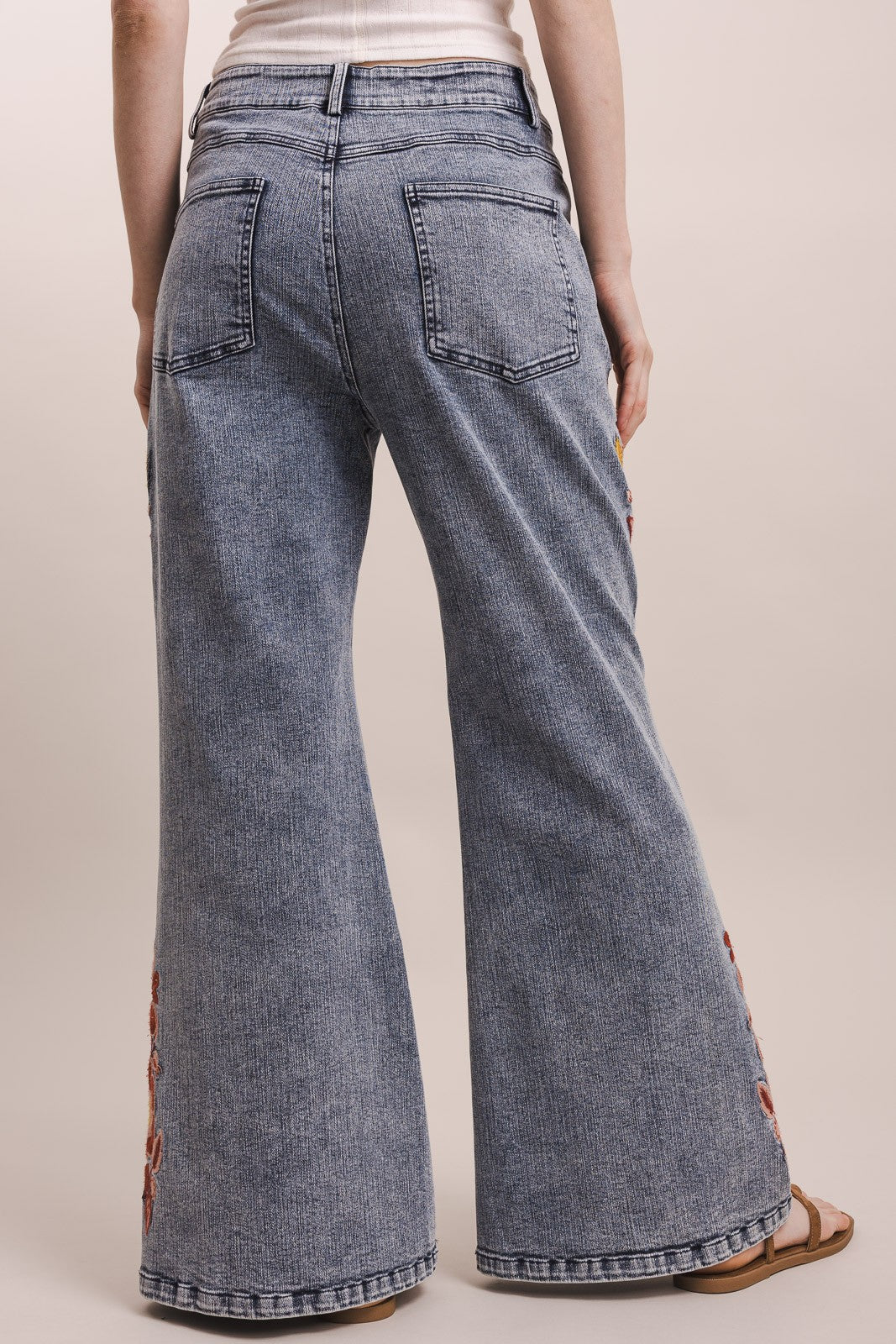 Wide-leg denim jeans in a washed blue-gray wash, shown from the back on a model. The jeans feature a high waist, back pockets, and a flared leg, with brown embroidered details running down the outer side of the legs.