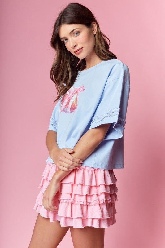 Light blue oversized tee with bow-wrapped football graphic and lace-trim sleeves, styled with a pink tiered ruffle mini skirt.