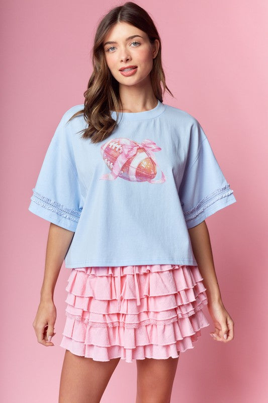 Powder blue oversized tee with a pink bow-wrapped football graphic at the chest; elbow-length sleeves trimmed with lace and a boxy, relaxed fit. Paired with a pink tiered ruffle skirt.