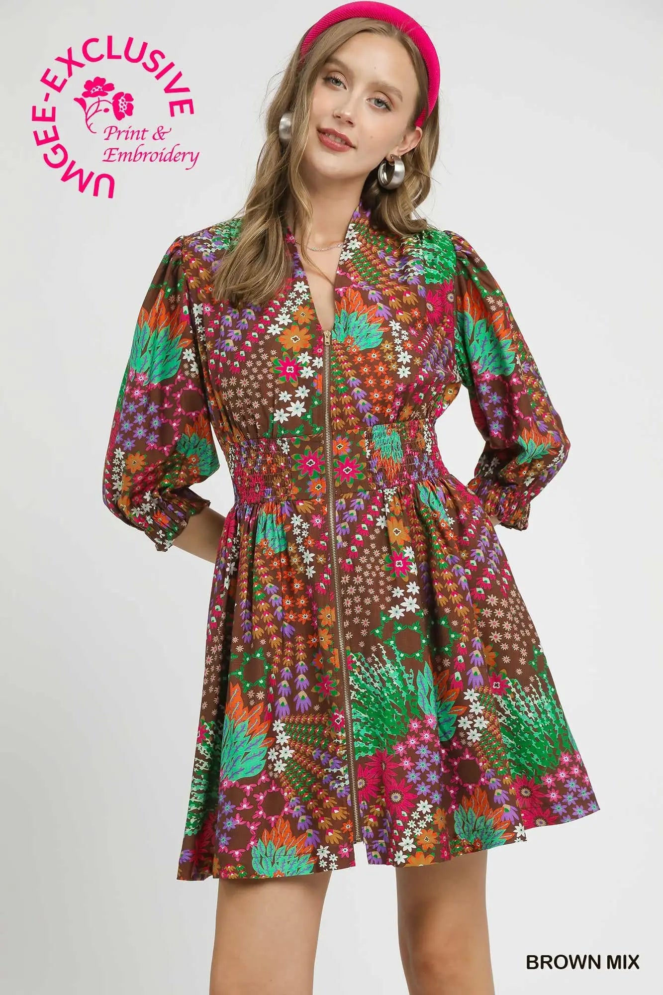 A woman modeling a short brown-based multicolor floral print dress with a stand collar, long sleeves, and a gathered waist. She is wearing a pink headband against a light gray studio background.