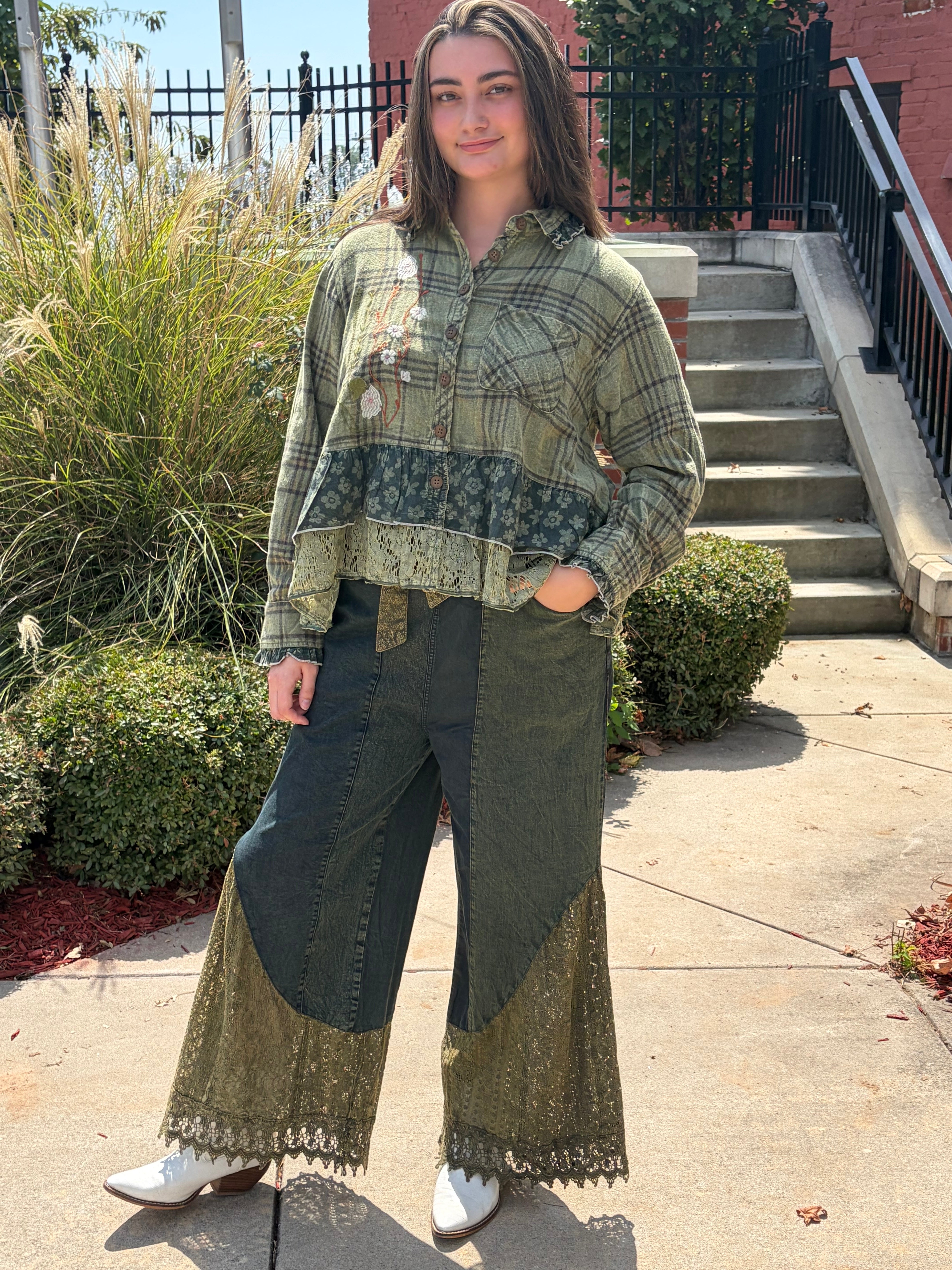A woman stands outdoors on a concrete walkway wearing a layered bohemian-style patchwork blouse and wide-leg patchwork pants. The outfit features olive green, beige, and dark gray sections with lace trim, paired with white boots.