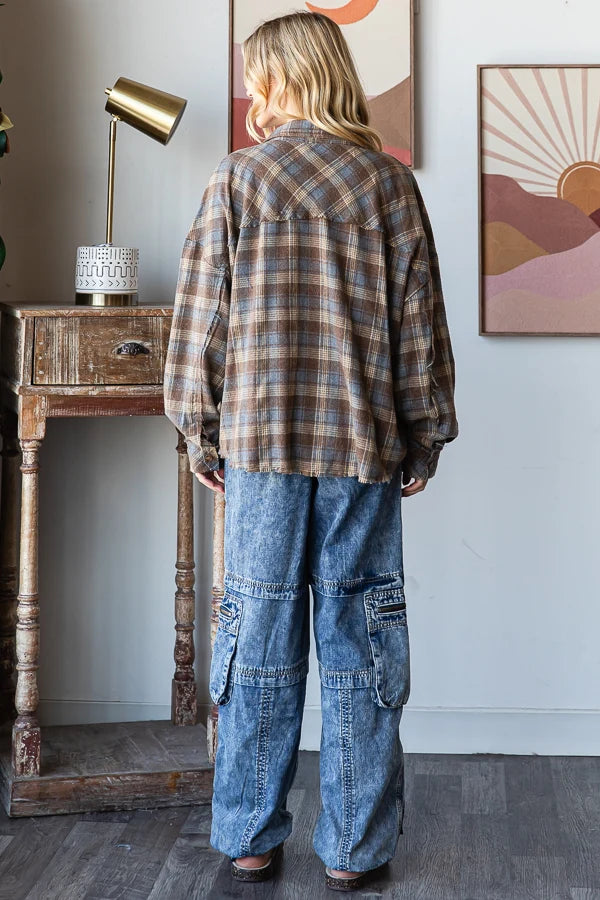 A person standing with their back to the camera, wearing a brown and beige plaid flannel shirt and wide-leg blue denim cargo pants in a styled room with a wooden side table and wall art.