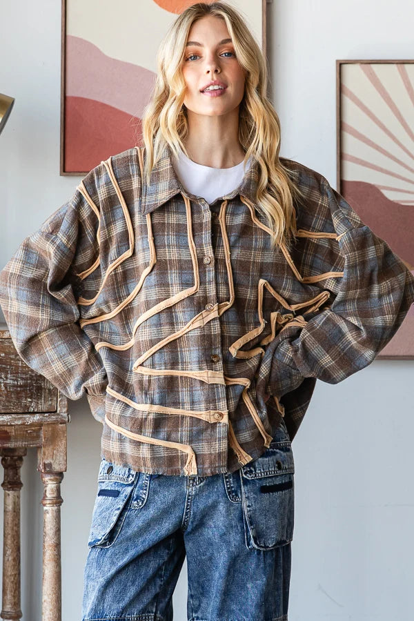 A woman wearing a brown and beige plaid button-up shirt jacket with decorative tan trim, styled with blue jeans over a white t-shirt, standing in a room with framed wall art.