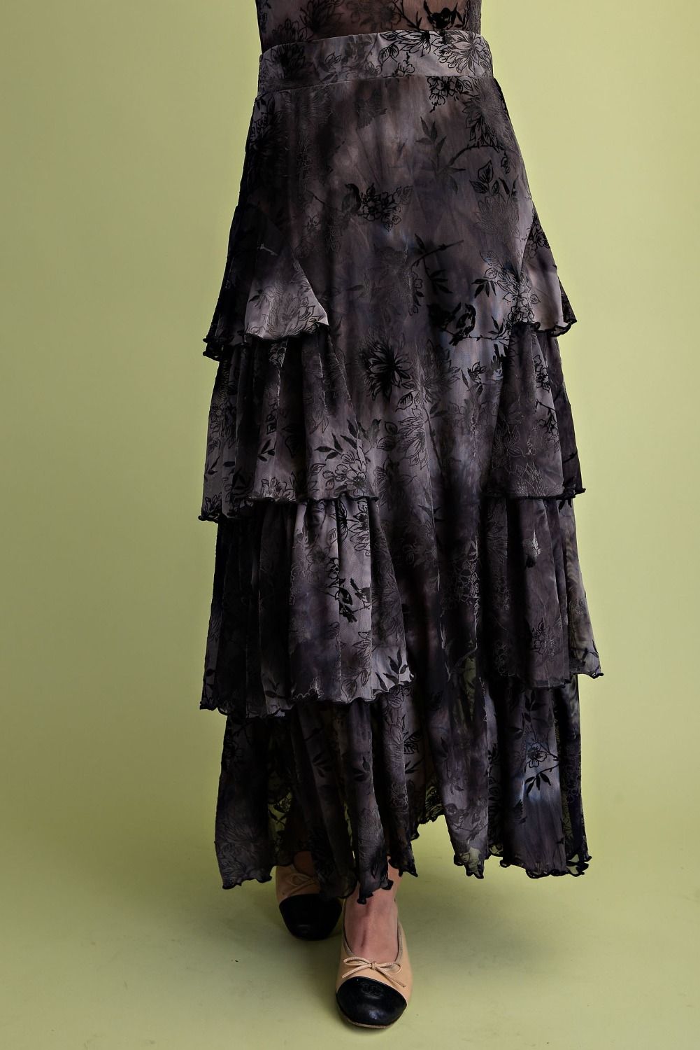 A long, tiered skirt in a dark charcoal gray and black floral pattern, worn by a model standing against a solid light olive green background.
