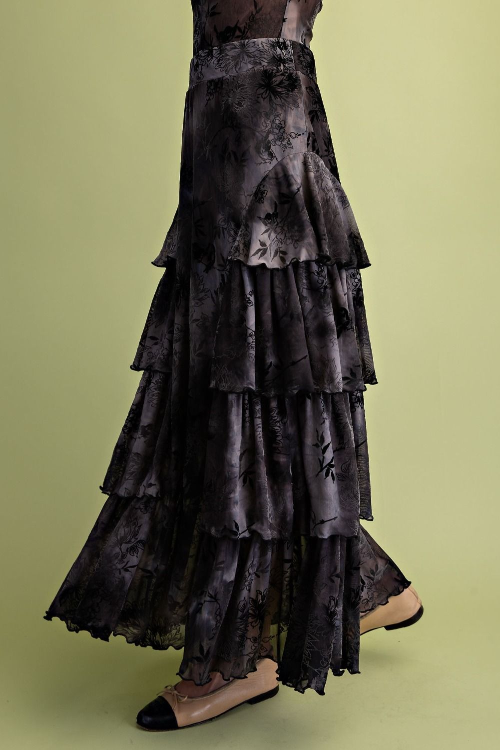 A long, tiered skirt with a dark floral print in black and gray, worn by a model with beige pointed-toe flats against a solid light olive green background.