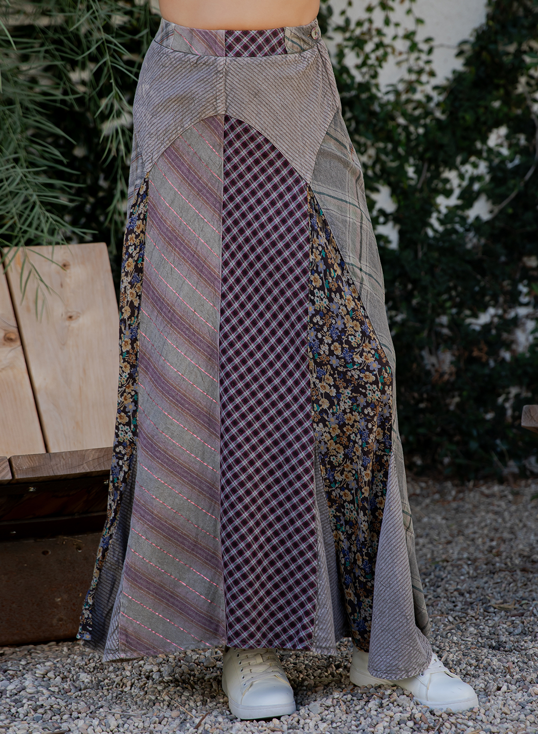 A long patchwork maxi skirt with multiple fabric panels including stripes, gingham, and floral prints in muted purple, gray, beige, and brown tones, shown on a model outdoors with white sneakers.