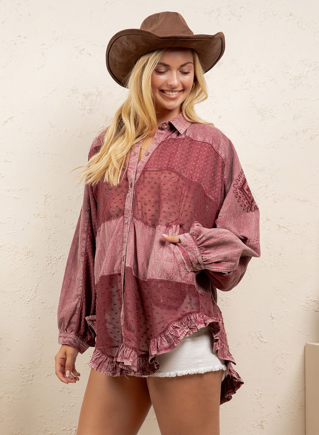 A woman wearing a dusty rose patchwork button-up shirt with ruffled edges, a brown cowboy hat, and white shorts, standing against a textured beige wall.